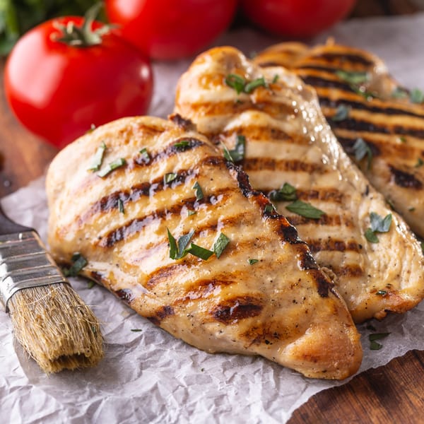 Grilled chicken breasts with grill marks and herbs, next to fresh tomatoes and a basting brush.