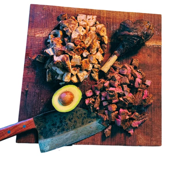 Chopped chicken, steak, a lamb shank, avocado, and a cleaver on a wooden board.